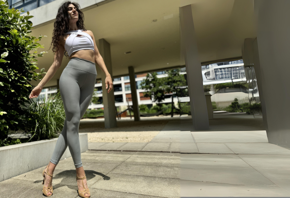 Eine Person in einem weißen Oberteil und grauen Leggings posiert selbstbewusst im Freien in der Nähe eines modernen Gebäudes.